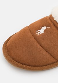 Pantoufle marron fabriquée en matériau doux semblable à du daim, avec un logo brodé blanc, un col rembourré et une semelle en caoutchouc texturé.