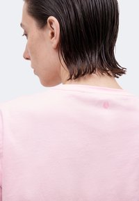 Άτομο με κοντά, βρεγμένα σκούρα μαλλιά φοράει ανοιχτό ροζ πουκάμισο, γυρισμένος ανάποδα, δείχνοντας το αριστερό προφίλ και το περίγραμμα του λαιμού σε απλό φόντο.