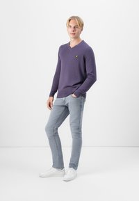 Pull v-neck violet en tissu doux, associé à un jean slim gris clair et des baskets blanches. Détail logo jaune sur le pull.