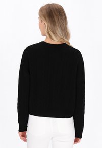 Zwarte kabelgebreide trui met lange mouwen en een ronde hals, met een cropped ontwerp en een gemeleerd patroon. Witte jeans zijn zichtbaar.