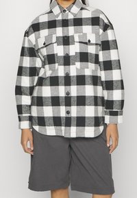 Black and white checkered flannel shirt with two chest pockets, button-up front, and long sleeves. Paired with dark gray shorts.