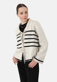 Cardigan lavorato a maglia beige con strisce orizzontali nere, aperto davanti con chiusura a fiocco e maniche ampie. Il tessuto ha una consistenza morbida.