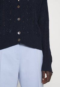 Main noire, portant un cardigan en tricot bleu marine avec des boutons, associé à un pantalon taille haute bleu clair, sur fond blanc.