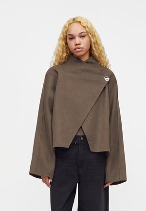 Jeune femme aux longs cheveux blonds bouclés portant une veste marron ample à manches larges et un pantalon noir taille haute.