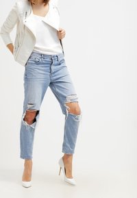 Jeans in denim azzurro chiaro strappati con bordi sfilacciati, indossati con una maglietta bianca e una giacca in pelle bianca testurizzata. Abbinati a tacchi alti bianchi.