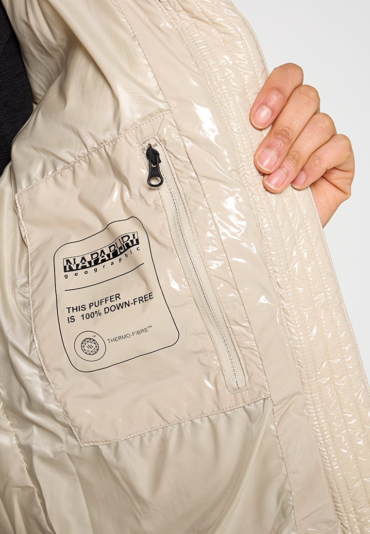 Beige Daunenjacke mit glänzender Textur. Verfügt über eine mit Reißverschluss gesicherte Tasche und ein Etikett mit der Aufschrift "100% daunenfrei" sowie dem Thermo-Fibre™-Markenlogo.