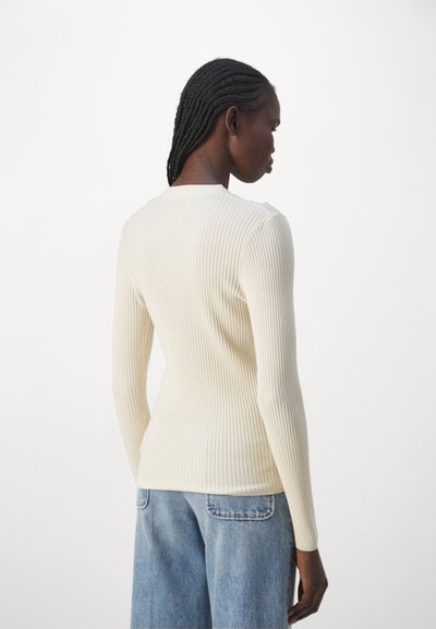 Langermet ribbestrikket topp i kremfarge, med rund hals og tettsittende silhuett, kombinert med lyseblå jeans. Strukturert strikkedetalj synlig.