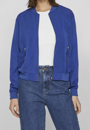 Vrouw draagt een blauwe jas met ritssluiting en zijzakken over een wit shirt en een blauwe jeans met hoge taille, tegen een effen achtergrond.