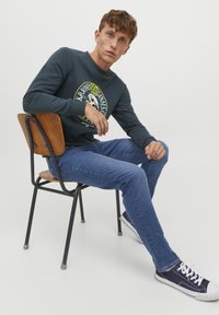 Maglietta a maniche lunghe blu scuro con stampa grafica, abbinata a jeans di lavaggio medio e sneakers blu navy, seduto su una sedia di legno contro uno sfondo bianco.