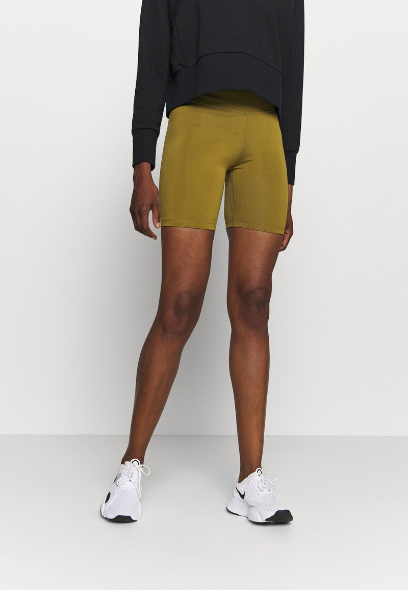Personne portant un short moulant vert olive, un haut à manches longues noir et des baskets blanches Nike, debout devant un fond clair uni.