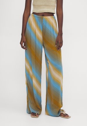 Kobieta w luźnych, wysokich spodniach z paskami ukośnymi w kolorach niebieskim, musztardowym i beżowym, zestawiona z beżowym krótkim topem i sandałami.
