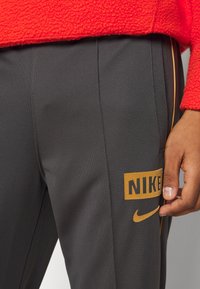Närbild av en hand som vilar på mörkgrå Nike-byxor med guldigt logo och sidostråk, person som bär en röd fleecejacka.