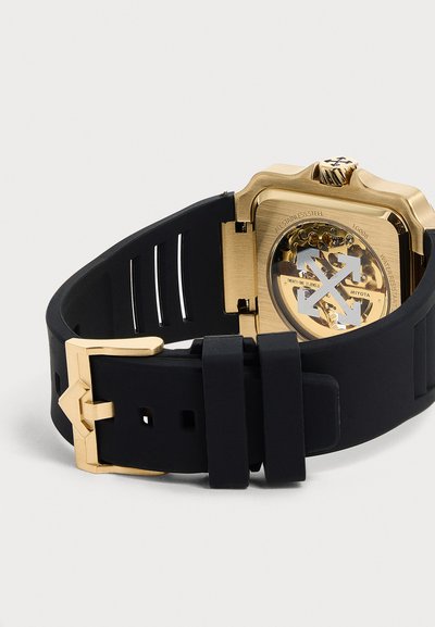 Boîtier de montre doré avec mouvement mécanique visible et bracelet en caoutchouc noir avec boucle dorée sur un fond neutre.
