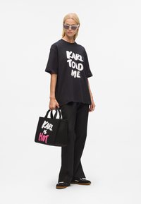 T-shirt noir surdimensionné avec le texte blanc "KARL M'A DIT", pantalon noir, portant un petit sac fourre-tout noir avec "KARL" et "HOT" en blanc et rose.