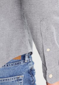 Zbliżenie na rękaw szarego wełnianego swetra na tle niebieskich dżinsów z widocznym przeszyciem i guzikami na mankiecie.