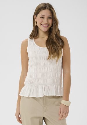 Noor naine pikkade pruunide juustega, kes kannab valget varrukateta topi ja beeže pükse, naeratab kuldsete rõngakõrvarõngaste ja kreemika käevõruga.