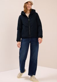Giacca puffer blu navy con cappuccio, chiusura a zip e tasche frontali. Abbinata a jeans a gamba larga e sneakers leggere, con una superficie testurizzata.