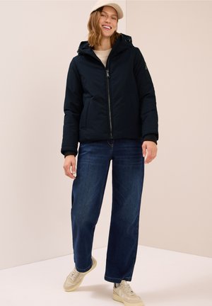 Marinblå puffjacka med huva, dragkedjestängning och framfickor. Kombineras med vida jeans och ljusa sneakers med en texturerad yta.