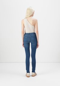 Top beige de un solo hombro combinado con jeans ajustados azules, con una cintura de altura media y bolsillos traseros. Zapatos de color claro completan el conjunto.