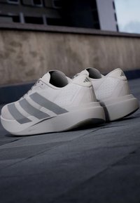 Λευκά και γκρι παπούτσια για τρέξιμο Adidas με μαξιλαρομένες σόλες τοποθετημένα σε πλακόστρωτη υπαίθρια επιφάνεια κοντά σε έναν τοίχο από σκυρόδεμα.