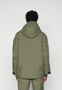 Olíva zöld, vízálló dzseki kapucnival, sima текстúrával, elülső varrásokkal és állítható mandzsettákkal. Csattok és cipzárok nem láthatók.