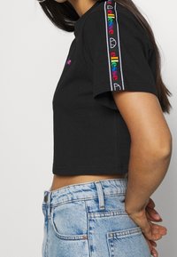 Camiseta negra corta con mangas cortas, que presenta un logo de "ellesse" multicolor en ambas mangas y una textura suave de algodón. Combinada con jeans de color azul claro.