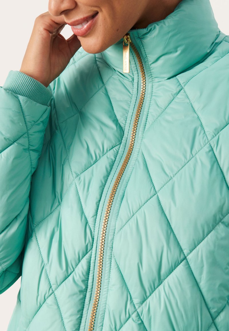 Chaqueta acolchada en azul claro que presenta un collar alto, cremallera dorada y puños acanalados, mostrando una textura suave y un patrón de diamantes.