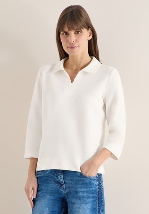 Femme aux cheveux bruns raides portant une chemise blanche à manches longues avec col et un jean bleu, debout contre un mur beige uni.
