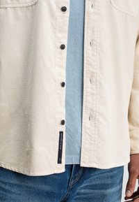 Beige overhemd met knopen en een gladde textuur met een subtiel nerfpatroon, gedragen over een lichtblauw T-shirt met ronde hals. Zwarte knopen en een labeldetail aan de zijkant.