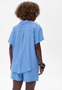 Lichtblauw overhemd met korte mouwen, een kraag en een plooi aan de achterkant, gecombineerd met bijpassende korte broek. Zachte textuur, ontspannen pasvorm en opgerolde mouwboorden.