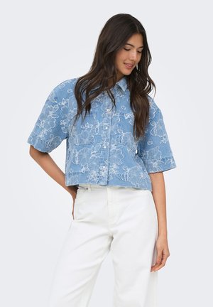 Mujer con cabello largo y oscuro que lleva una camisa azul con flores de manga corta abotonada y pantalones blancos de cintura alta, posando con una mano en la cadera.