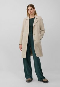 Vrouw die staat en een beige geruite lange jas draagt over een donkergroen top en bijpassende wijde broek met beige schoenen met een chunky zool.