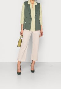 Gilet vert croisé porté sur un haut vert clair, associé à un pantalon court beige clair et des talons vert foncé. Petit sac à main transparent.