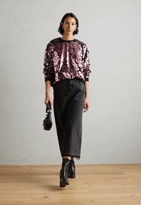 Suéter rosa con lentejuelas y puños negros, combinado con una falda de mezclilla negra de cintura alta y botas negras hasta el tobillo, sosteniendo un pequeño bolso negro.