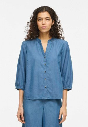 Kobieta o kręconych włosach, ubrana w niebieską bluzkę na guziki z rękawami do łokci oraz pasujące niebieskie spodnie, stojąca na białym tle.