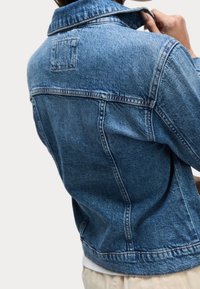 Svetle modrá džínová bunda s klasickým střihem, která má špičatý límeček, přední yoke a viditelné švy. Textura je hladká s mírným vyblednutím.