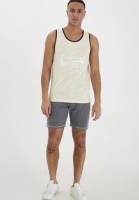 Lysegul top med grafisk print, marineblå kant, parret med mørkegrå shorts og hvide sneakers på en neutral baggrund.