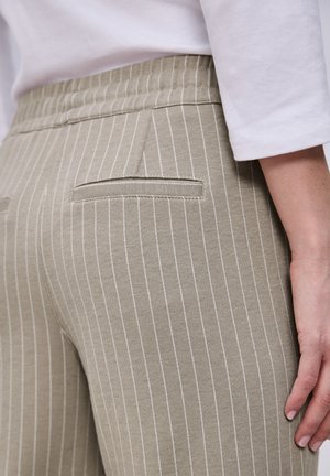 Close-up af beige bukser med striber og baglommer med kant, båret med en hvid bluse med lange ærmer, hånd hvilende nær hoften.