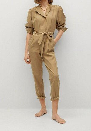 Kvinde står barfodet iført en tan jumpsuit med opkørte ærmer og et stofbælte bundet om taljen mod en ensartet baggrund.