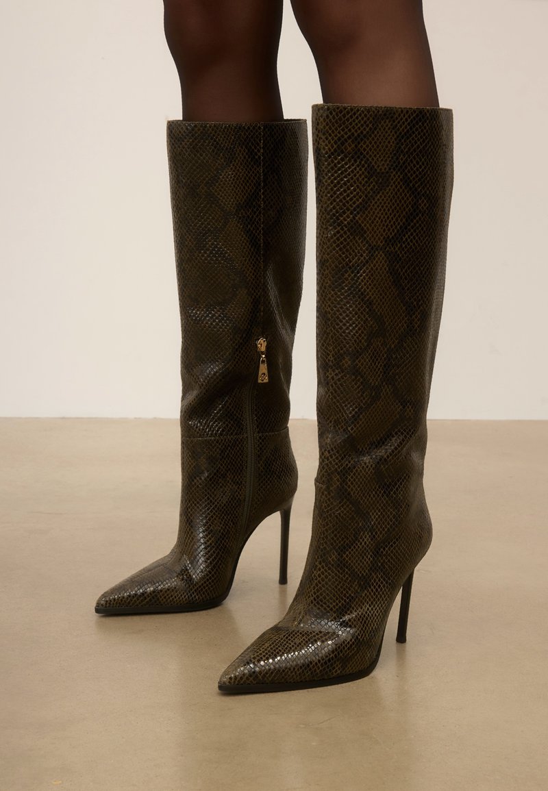 Bottes stilettos à talons aiguilles à bout pointu montant jusqu’au genou avec un motif peau de serpent marron, portées sur des jambes à peau foncée sur un fond uni de sol et mur.