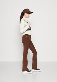 Bruine fluwelen flared broek, witte hoge sneakers, oversized romige sweater en een bruine pet met een logo, terwijl een kleine zwarte handtas wordt vastgehouden.