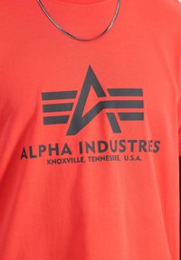 Camiseta de algodón roja de manga corta con un gran logotipo gráfico en negro y el texto "ALPHA INDUSTRIES, KNOXVILLE, TENNESSEE, U.S.A." en la parte frontal.