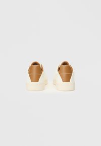 B baskets blanches avec un contrefort de talon en cuir beige, présentant un dessus lisse, un bout rond et un design simple. La semelle en caoutchouc est de couleur claire.