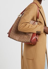 Sac en tissu à motif marron avec des accents en cuir rouge, de forme structurée, avec des éléments en métal doré et une bandouli ère amovible.