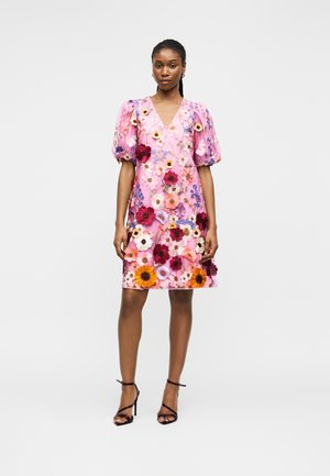 Kvinde, der står iført en pink V-hals kjole med pufærmer og farverig 3D blomsterbroderi, kombineret med sorte højhælede sandaler.