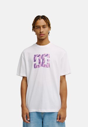 Junger Mann mit kurzen Zöpfen trägt ein weißes T-Shirt mit lila gemustertem Logo und blaue Jeans, steht vor einem schlichten, hellen Hintergrund.