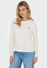Hvid langærmet t-shirt i bomuld med rund halsudskæring og en lille rød hjertebrodering på brystet, parret med blå jeans.