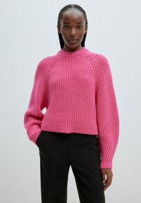 Pull en tricot rose avec un col montant, des épaules descendues et une texture côtelée; associé à un pantalon noir, au design court.