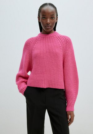 Gebreide roze trui met hoge nek, drop shoulders en ribbelstructuur; gecombineerd met zwarte broeken met een cropped ontwerp.
