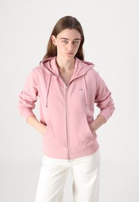 Młoda kobieta ubrana w różową bluzę z kapturem zapinaną na zamek z kieszeniami, białe spodnie, stoi z rękami w kieszeniach na jednolitym jasnym tle.